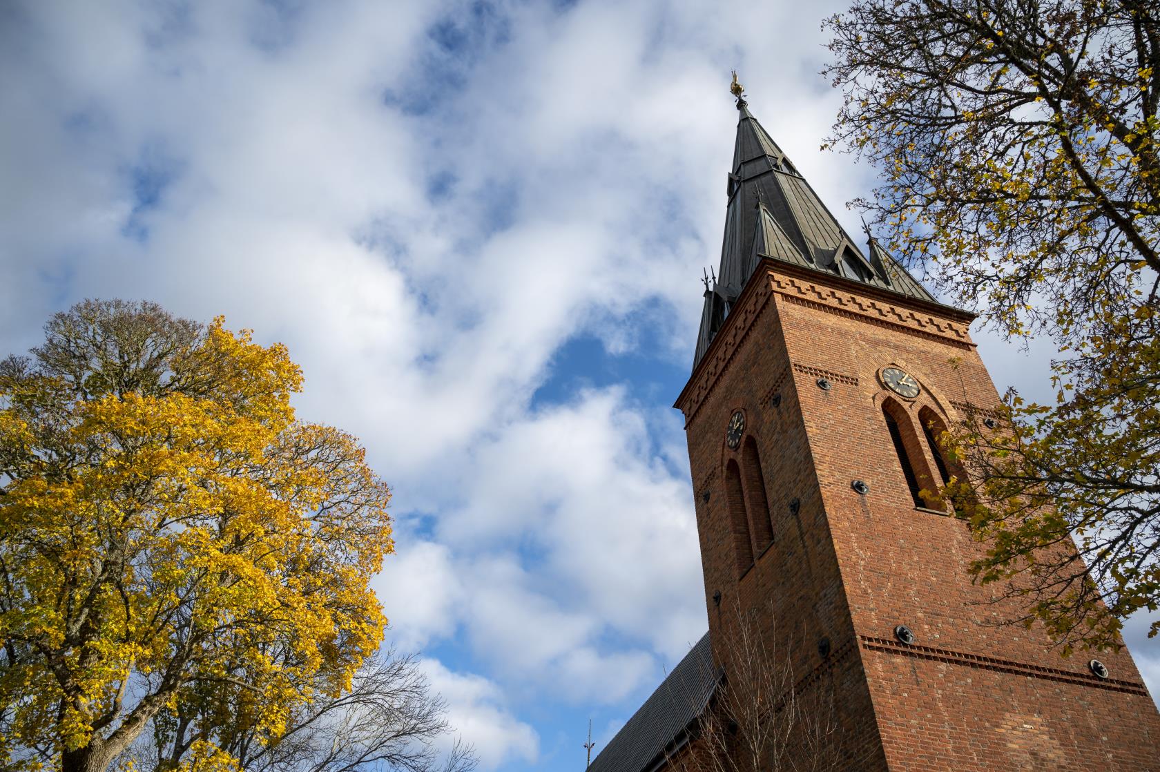 Ett kyrktorn med tegelfasad mot en blå och molnig himmel. Höstträd i förgrunden.