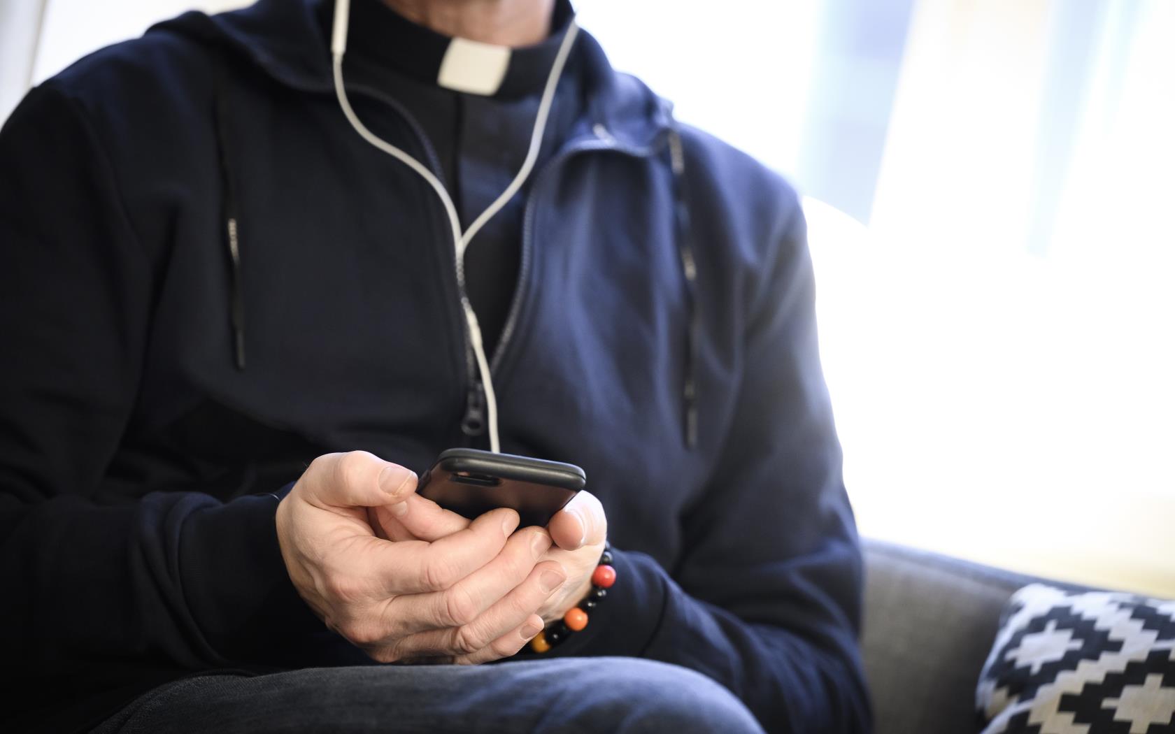 En manlig präst sitter i en soffa med en mobiltelefon i handen och hörlurar i öronen.