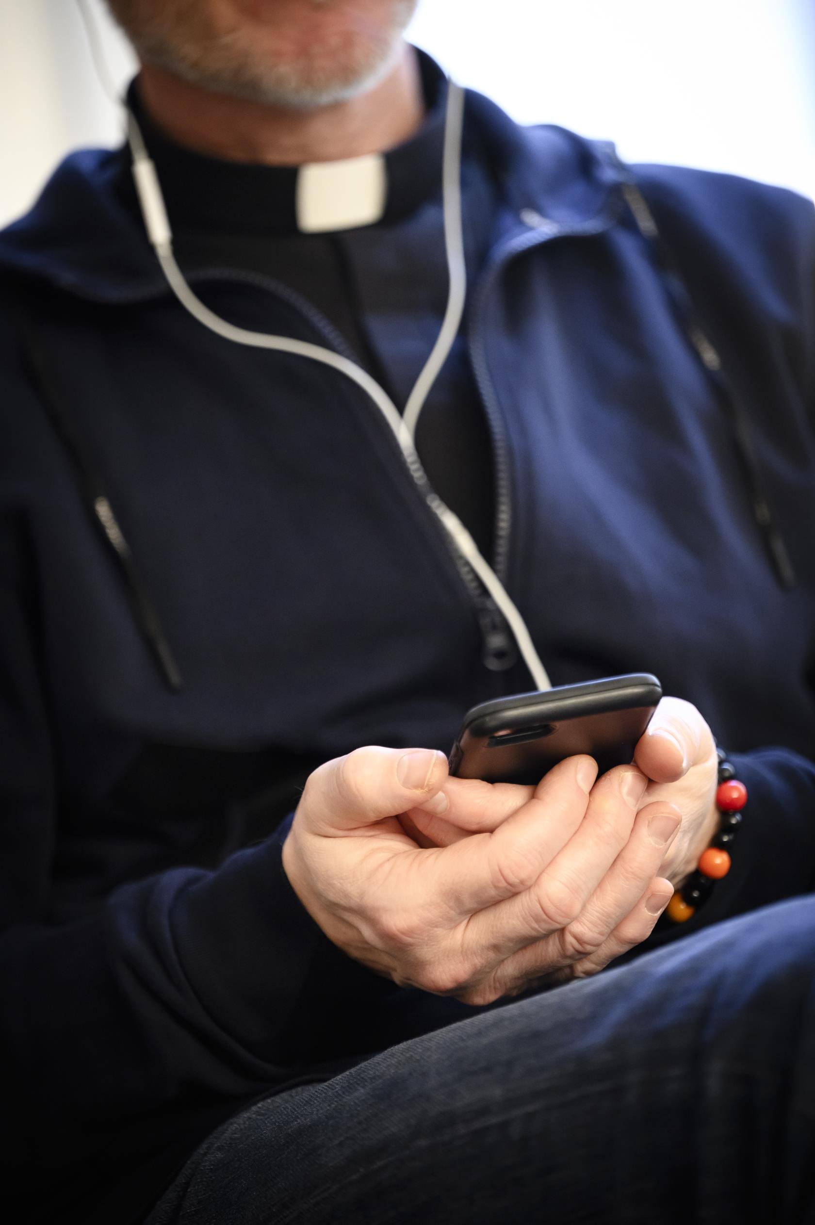 En manlig präst sitter i en soffa med en mobiltelefon i handen och hörlurar i öronen.