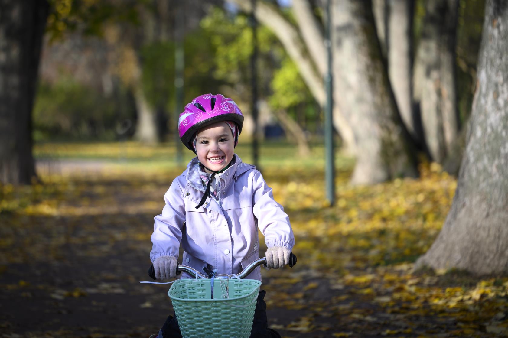 Ett barn med rosa hjälm cyklar genom en park full av höstlöv på marken.