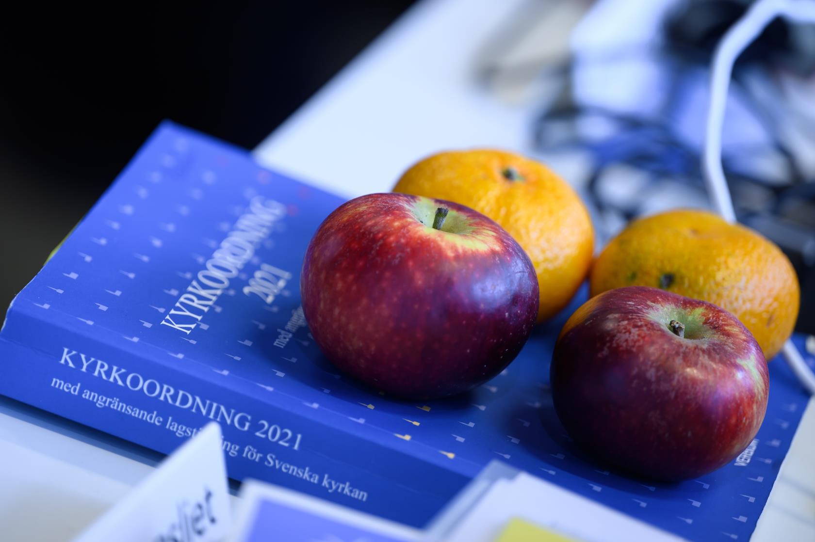 Två äpplen och två clementiner ligger på en blå bok med titeln Kyrkoordning 2021.