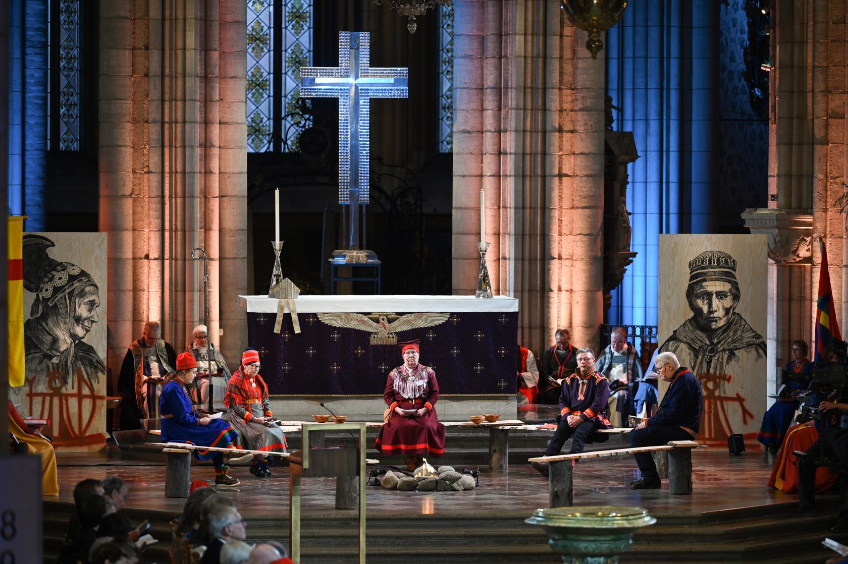 Fem personer i samedräkt sitter på grovhuggna bänkar runt en lägereld framför Uppsala domkyrkas altare.