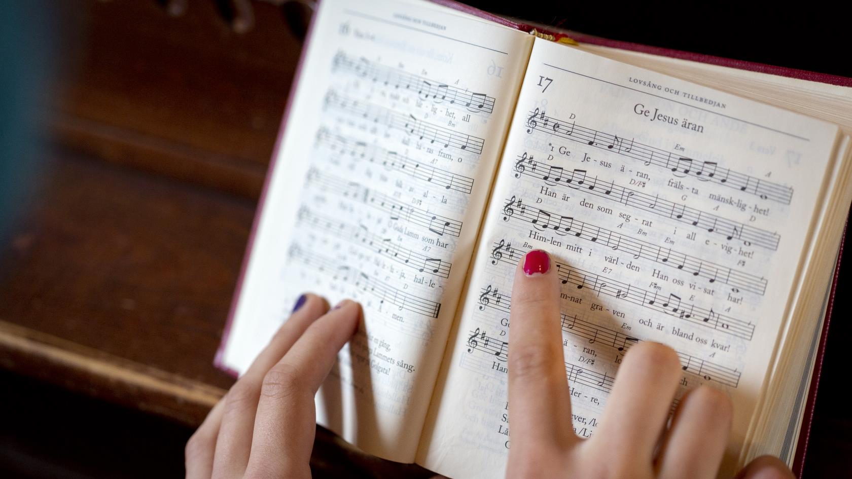 En person med målade naglar följer med sitt pekfinger melodin i en uppslagen psalmbok.