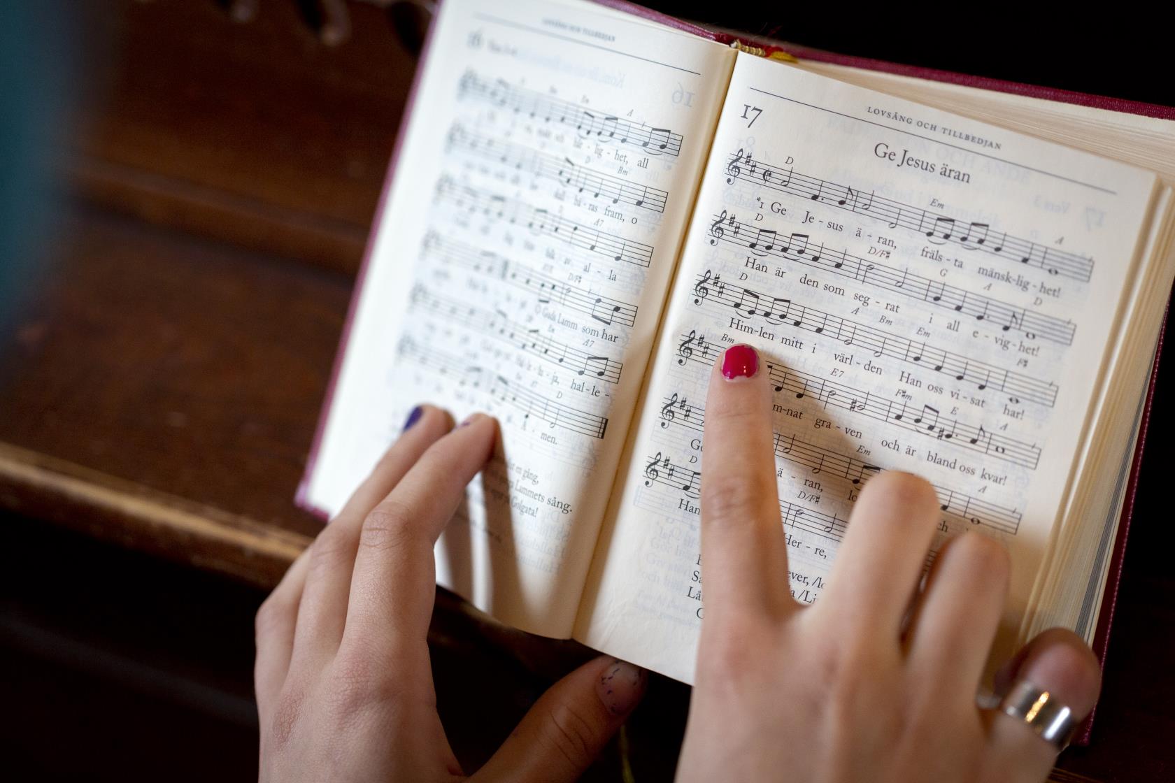 En person med målade naglar följer med sitt pekfinger melodin i en uppslagen psalmbok.