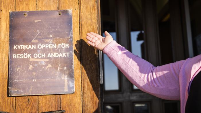Någon håller upp en arm mot en skylt på en kyrkport, med texten Kyrkan öppen för andakt.