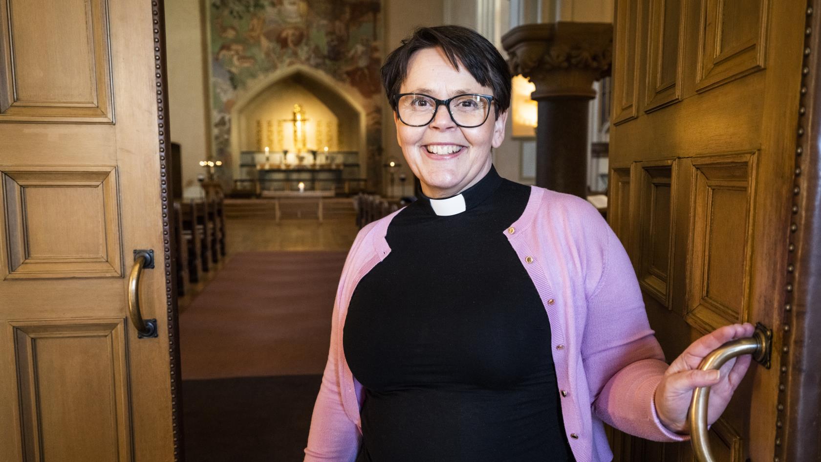 En kvinnlig präst i svart och med rosa kofta håller upp porten till hennes kyrka.