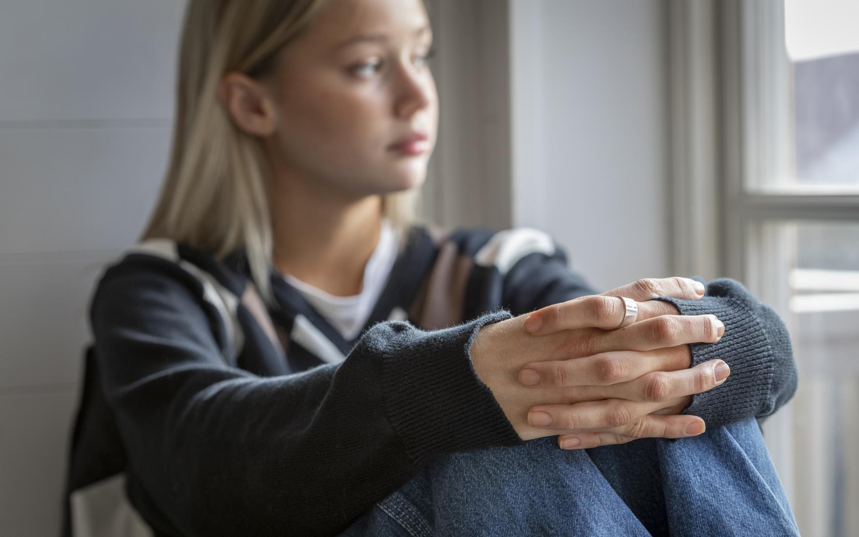 En tonårstjej sitter vid ett fönster med händerna om knäna.