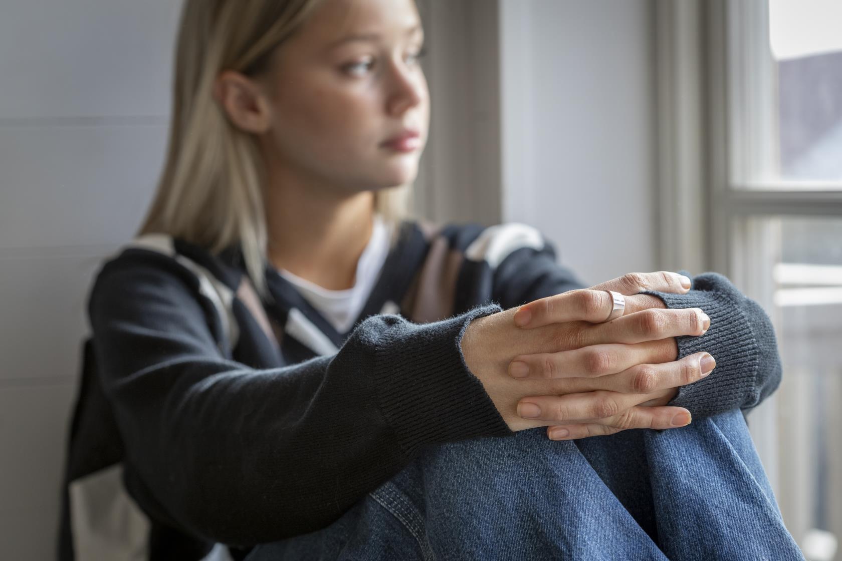 En tonårstjej sitter vid ett fönster med händerna om knäna.