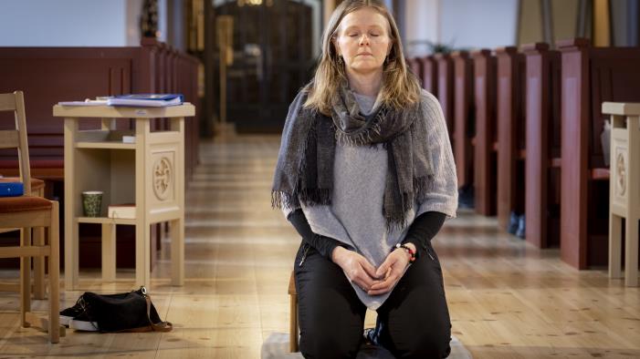 En kvinna sitter på en bönepall på en kyrkas golv, med knäppta händer och slutna ögon.