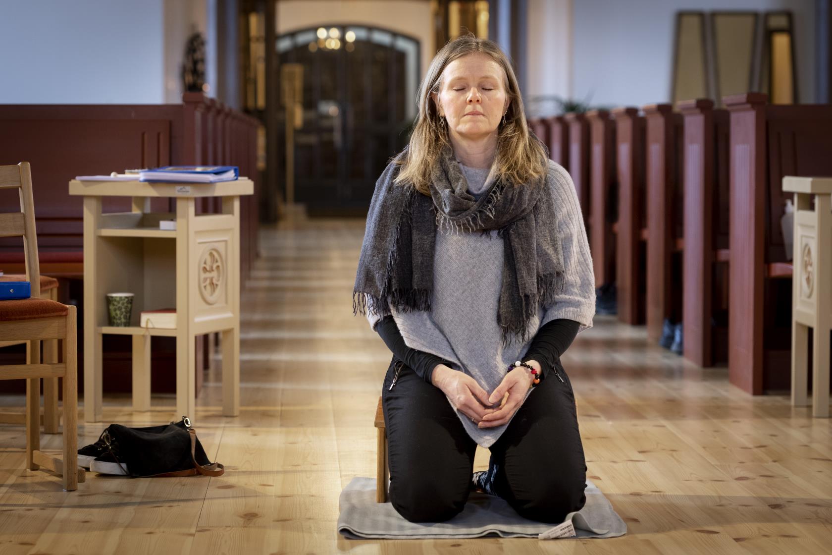 En kvinna sitter på en bönepall på en kyrkas golv, med knäppta händer och slutna ögon.