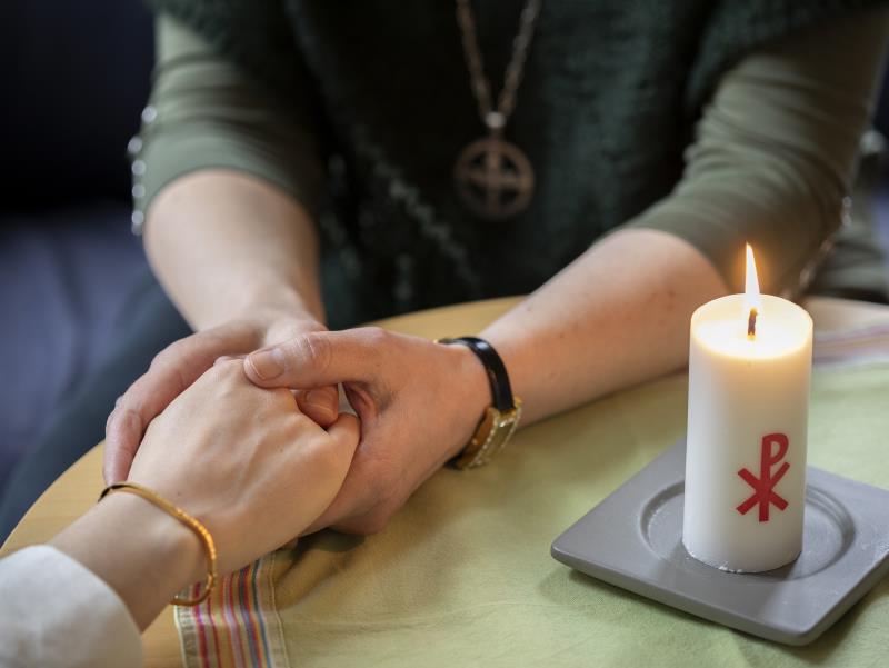 En diakon håller en annans hand i sina händer. Ett stearinljus med kristusmonogram brinner på bordet mellan dem.