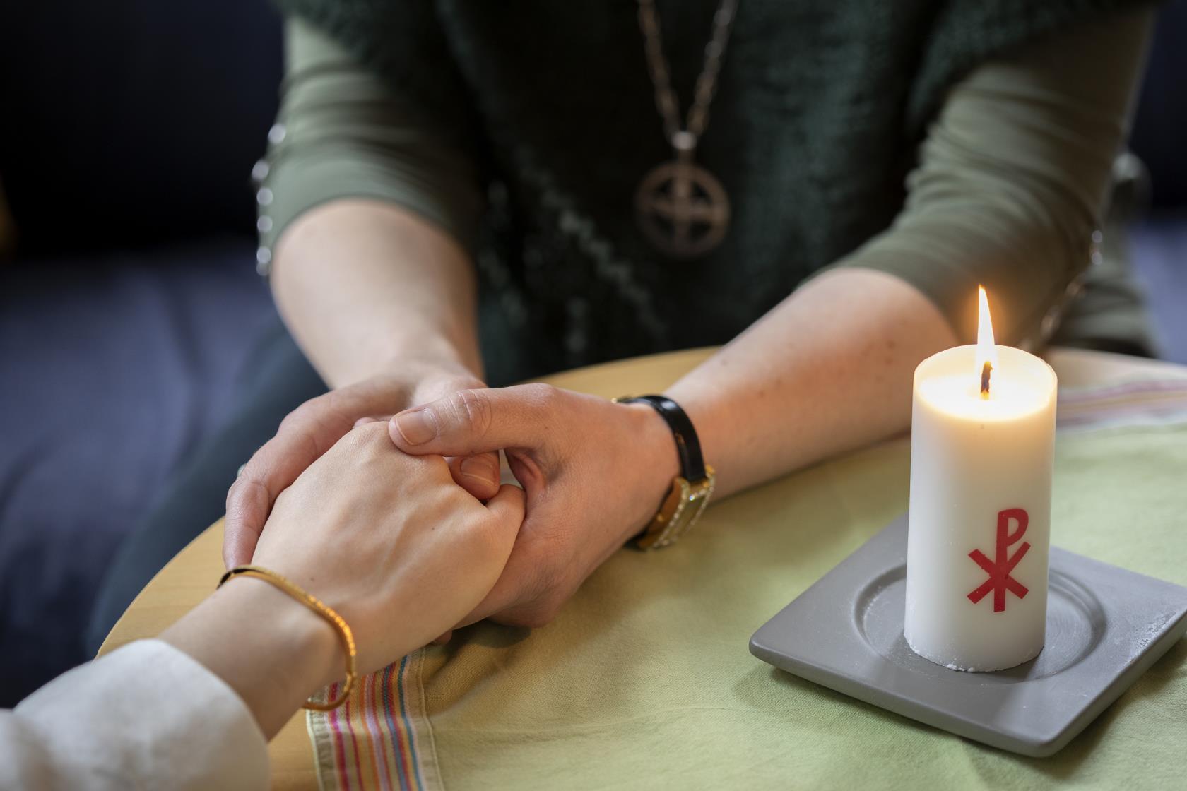 En diakon håller en annans hand i sina händer. Ett stearinljus med kristusmonogram brinner på bordet mellan dem.