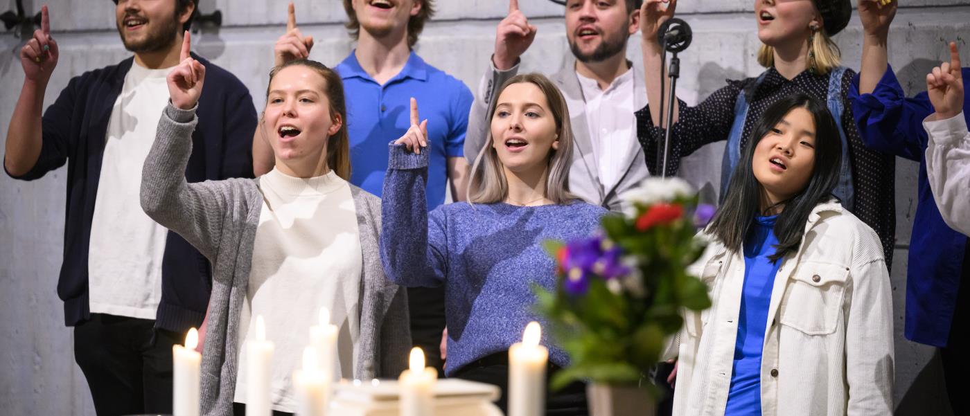 En blandad ungdomskör sjunger i en kyrka. De håller upp sina pekfingrar.