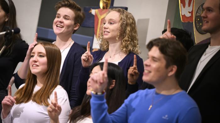 En blandad ungdomskör sjunger i en kyrka. De håller upp sina pekfingrar.