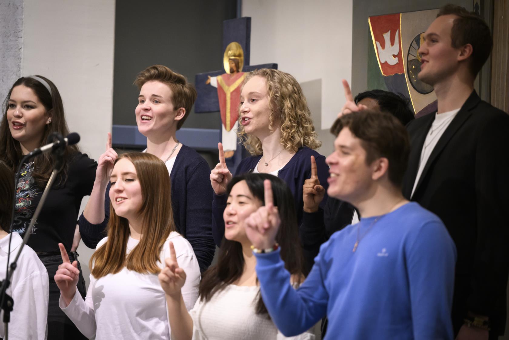 En blandad ungdomskör sjunger i en kyrka. De håller upp sina pekfingrar.