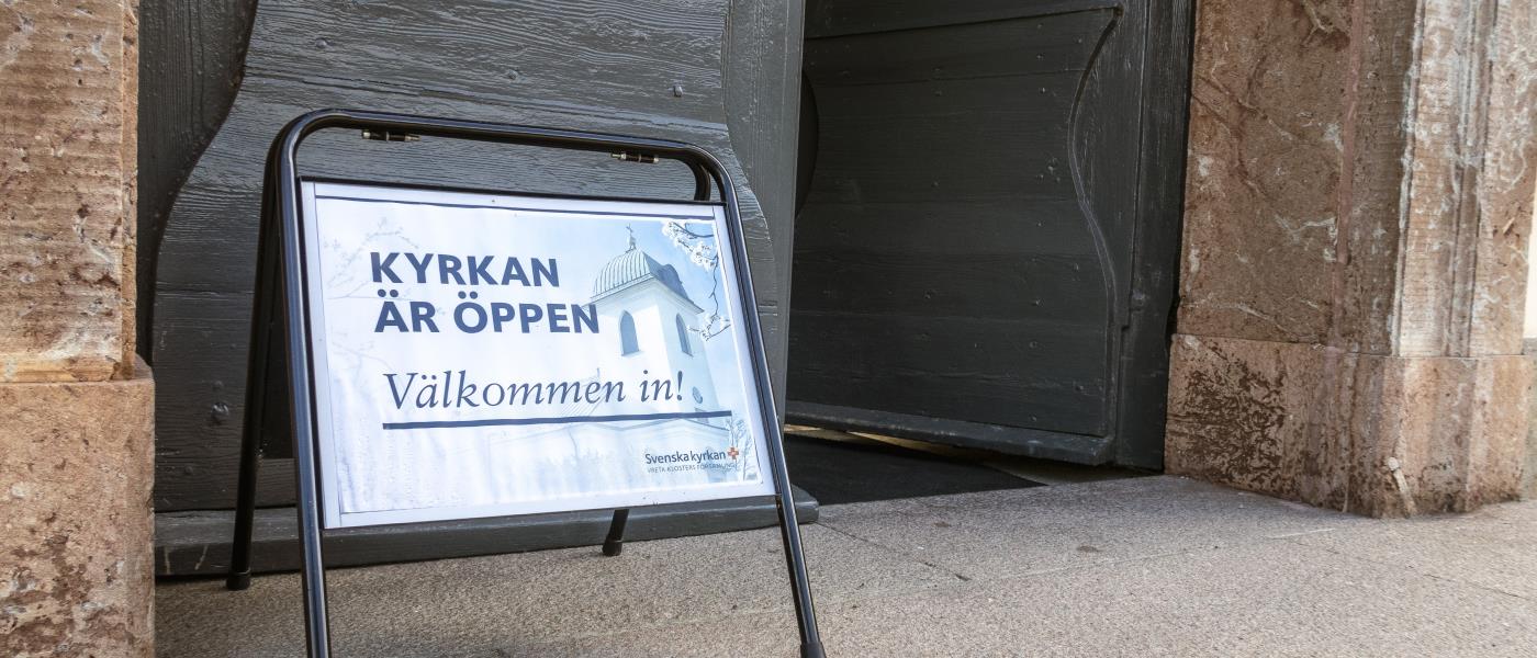 En skylt med texten Kyrkan är öppen välkommen in, står utanför en port.