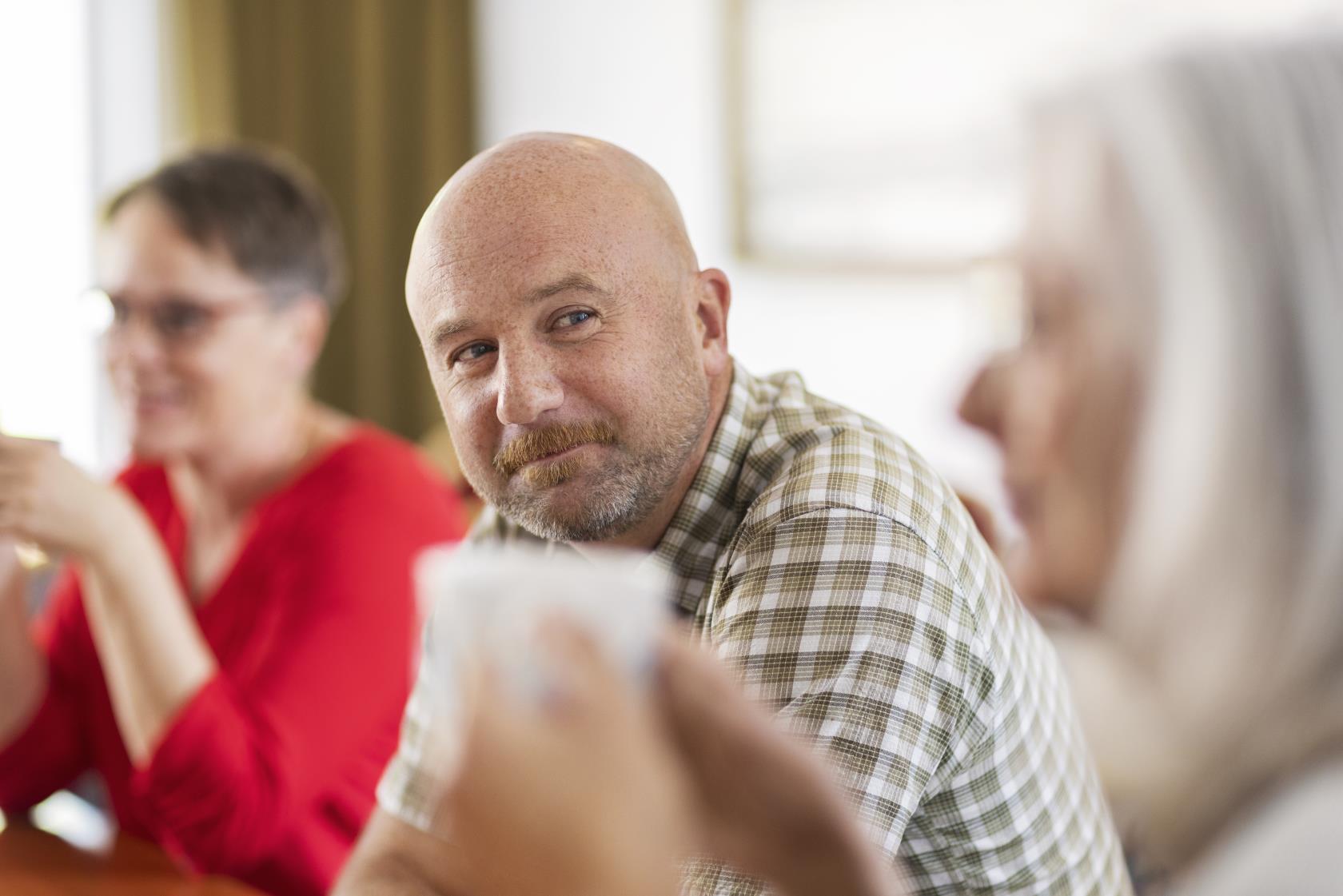 En man i rutig skjorta ler mot kvinnan bredvid. En kvinna med röd blus syns suddigt i bakgrunden.