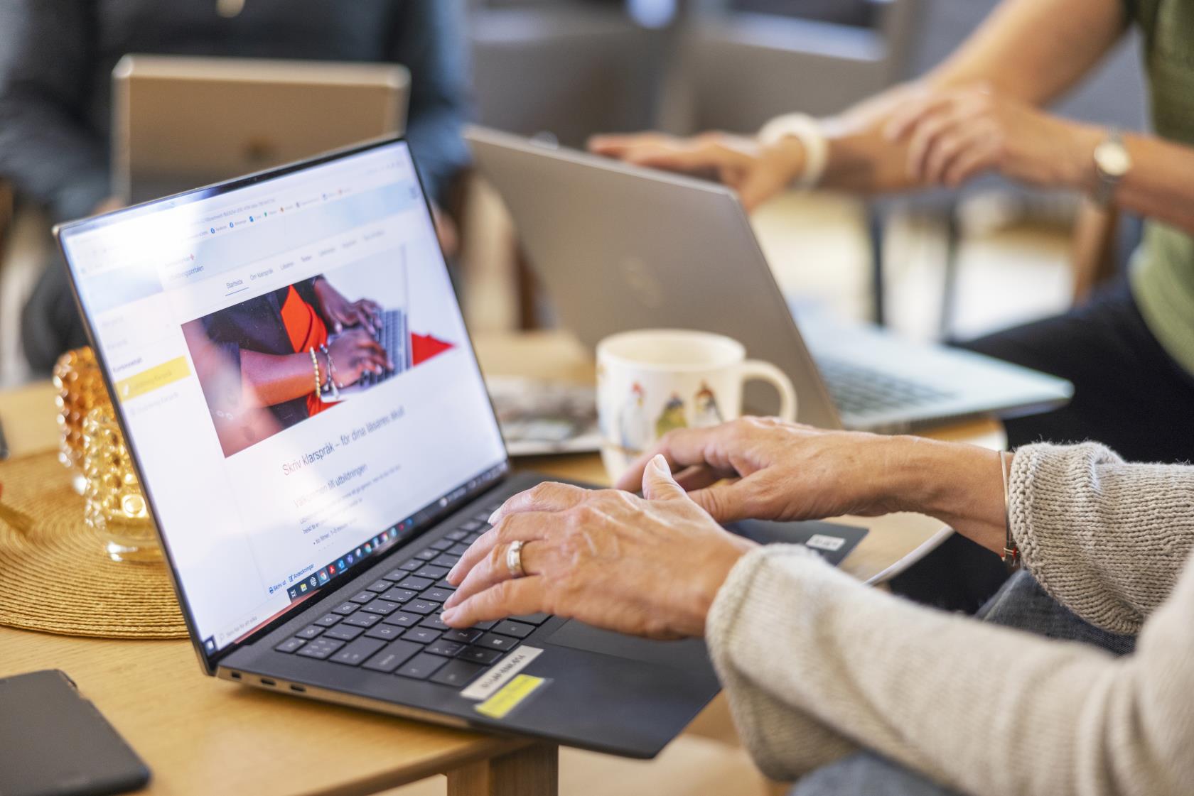 Någon skriver på en laptop. Två personer syns suddigt i bakgrunden.