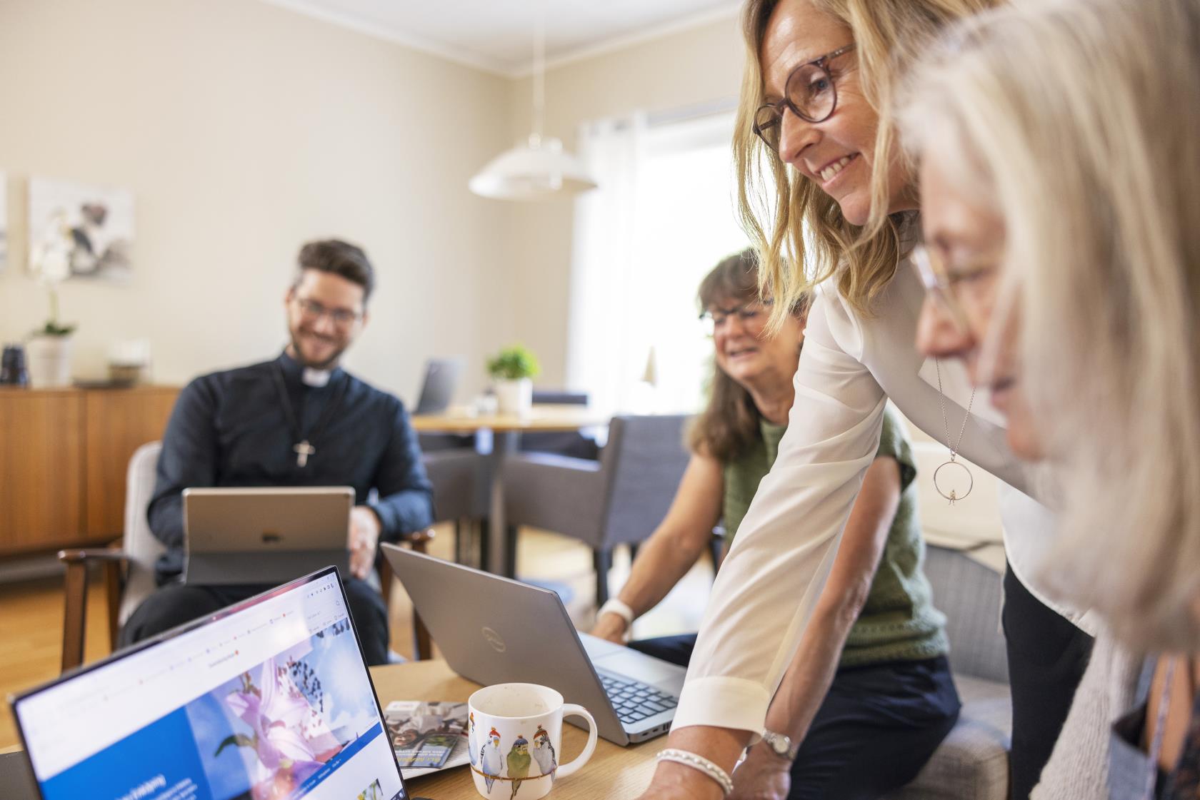 Två kvinnor tittar på en datorskärm. En manlig präst och en kvinna syns suddigt i bakgrunden.