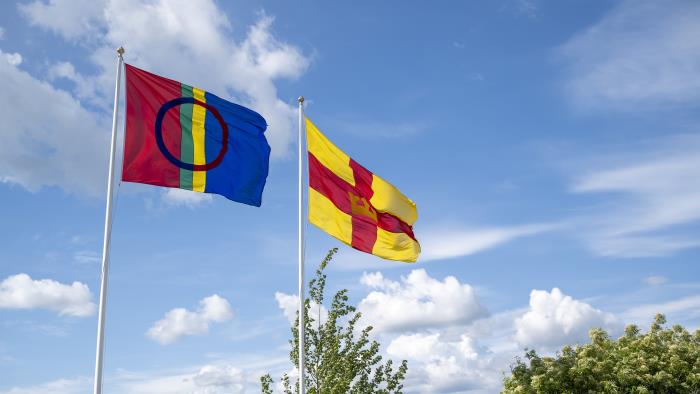 Två flaggstänger med den samiska flaggan och Svenska kyrkans flagga mot en blå himmel.
