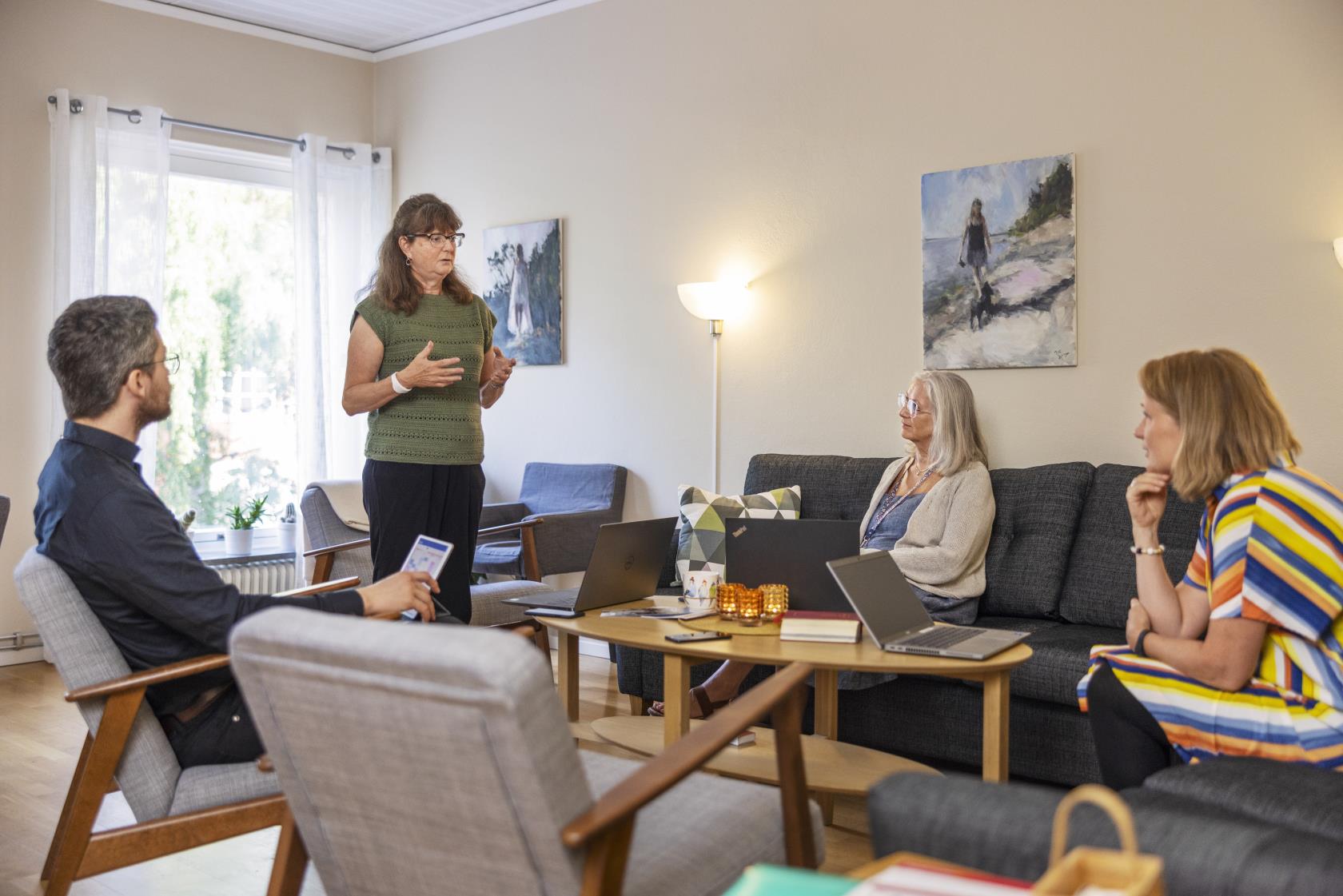 Tre personer sitter i en soffgrupp och lyssnar på en kvinna som står upp framför dem och pratar.