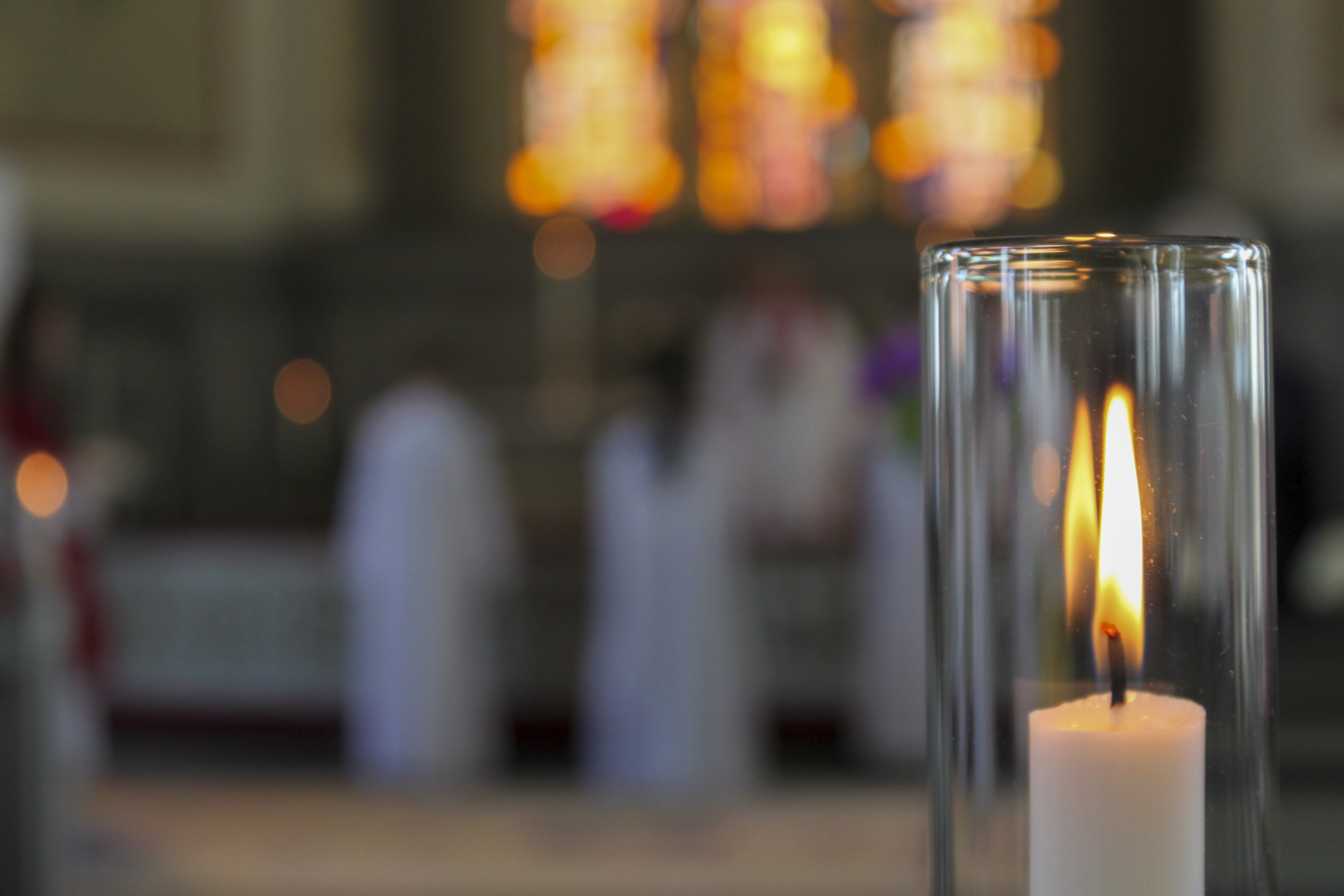 Närbild på ett brinnande ljus i en ljusstake med stormglas i en kyrka. I bakgrunden syns några personer vid altaret.