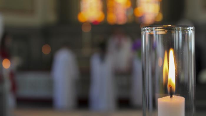 Närbild på ett brinnande ljus i en ljusstake med stormglas i en kyrka. I bakgrunden syns några personer vid altaret.