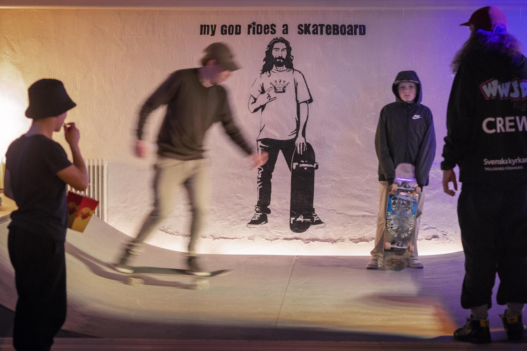 Ungdomar åker skateboard på en ramp. På väggen bakom är en målning och texten My god rides a skateboard.