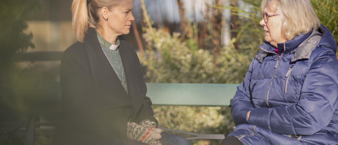 En kvinnlig diakon och en äldre kvinna sitter utomhus på en bänk och samtalar.