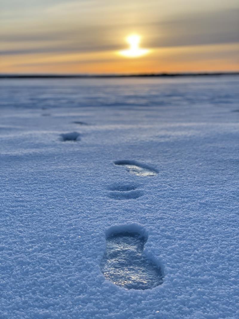 Ett par skoavtryck i snön. Solen går ner i bakgrunden.