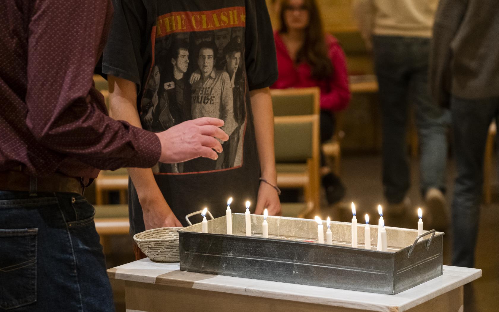 Några personer i en kyrka står samlade runt en liten plåtlåda fylld med smala brinnande stearinljus.