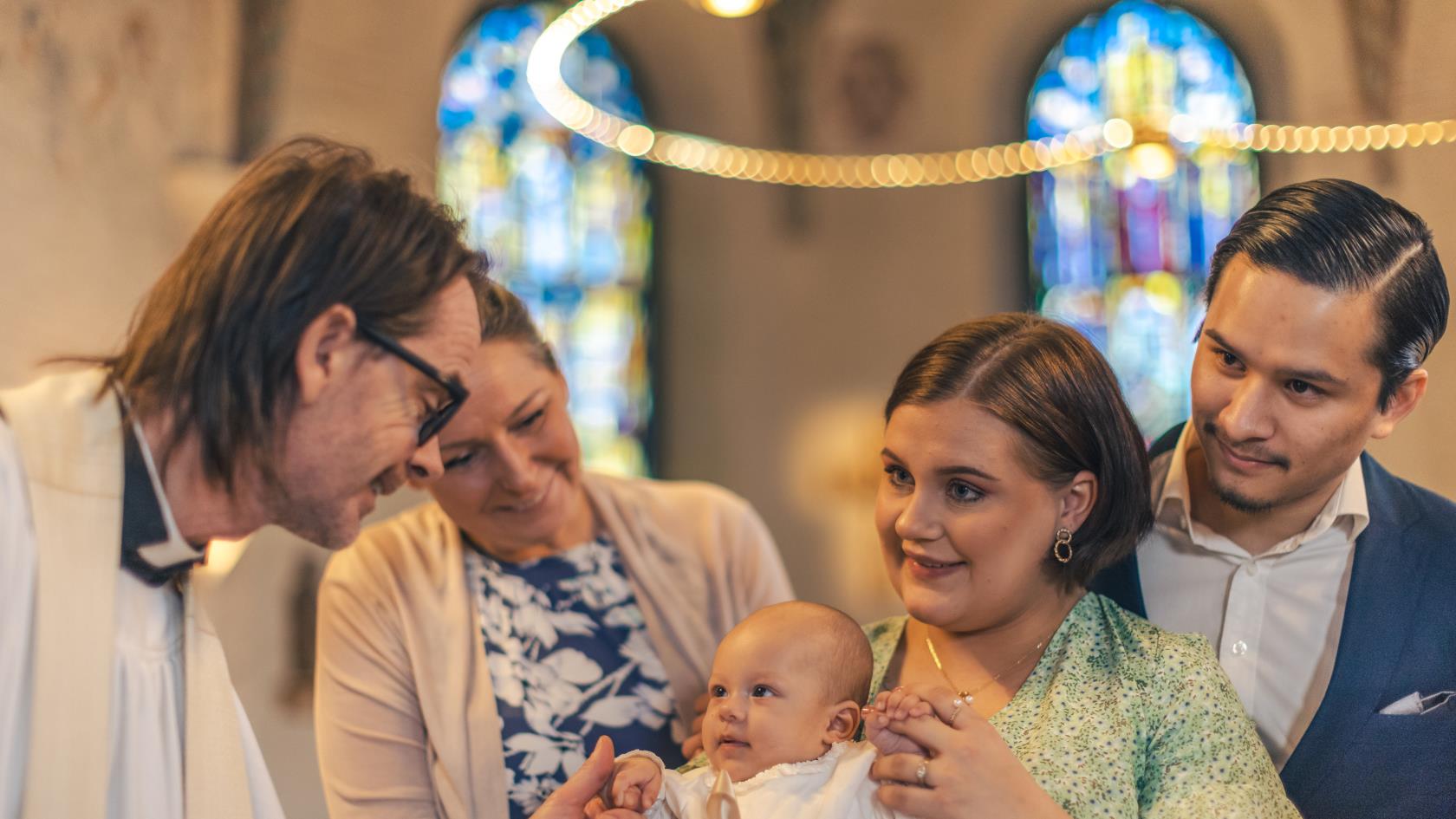 Ett dop i en kyrka. En kvinna med sin man bakom sig, håller barnet i famnen. En manlig präst håller barnets hand.