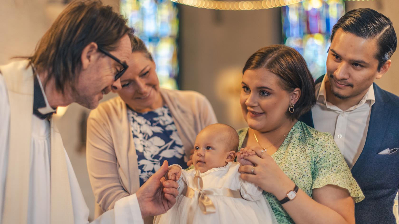 Ett dop i en kyrka. En kvinna med sin man bakom sig, håller barnet i famnen. En manlig präst håller barnets hand.