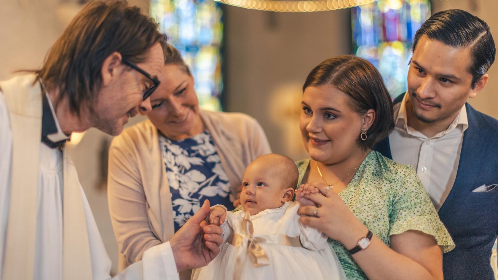 Ett dop i en kyrka. En kvinna med sin man bakom sig, håller barnet i famnen. En manlig präst håller barnets hand.