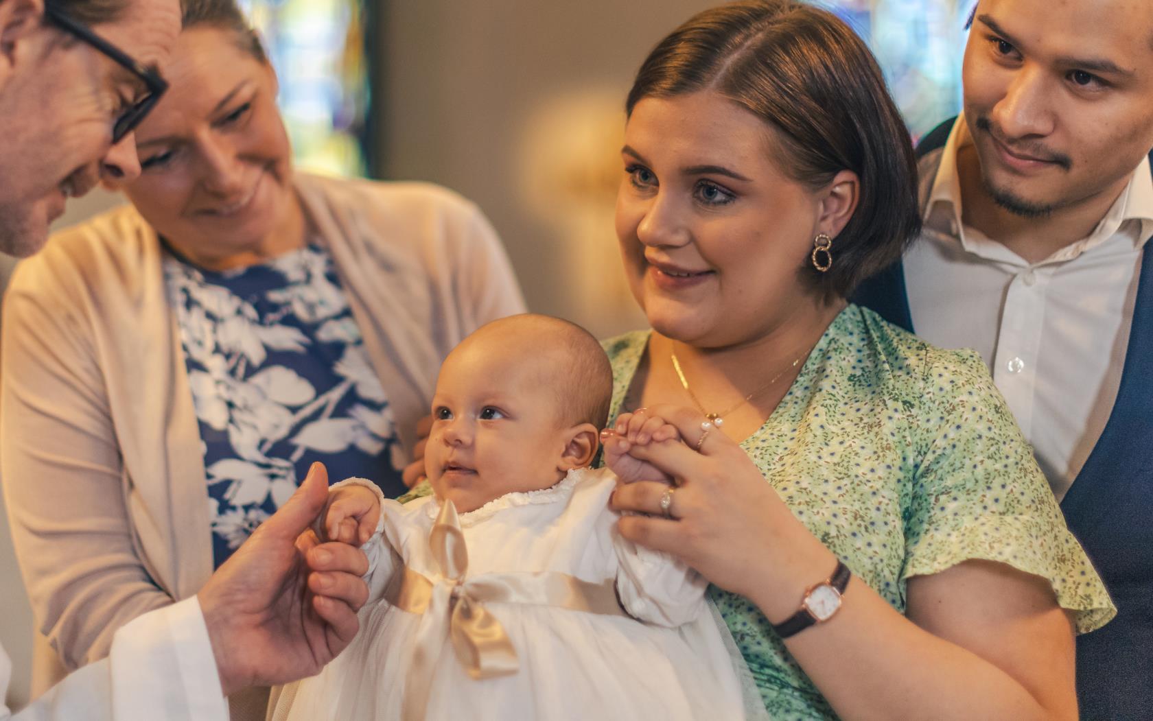 Ett dop i en kyrka. En kvinna med sin man bakom sig, håller barnet i famnen. En manlig präst håller barnets hand.