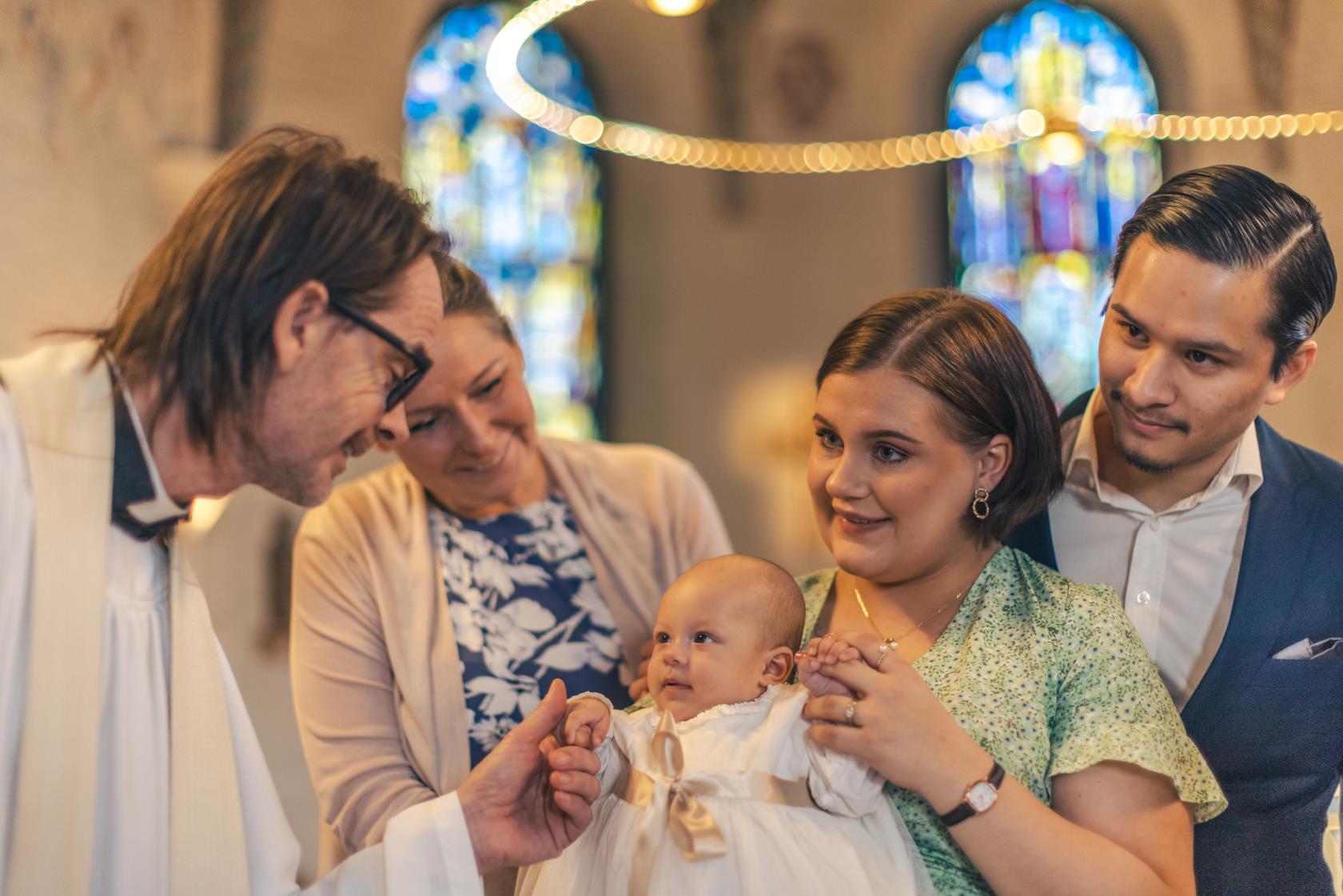 Ett dop i en kyrka. En kvinna med sin man bakom sig, håller barnet i famnen. En manlig präst håller barnets hand.