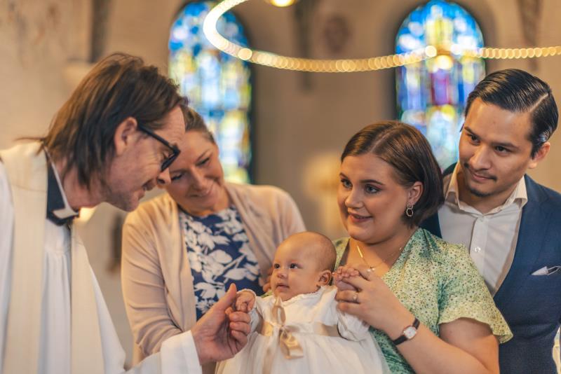 Ett dop i en kyrka. En kvinna med sin man bakom sig, håller barnet i famnen. En manlig präst håller barnets hand.