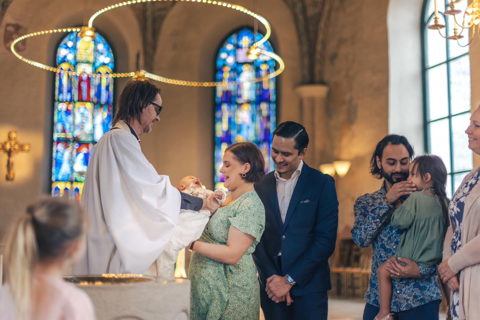 Ett dop i en kyrka. En manlig präst tar bebisen ur famnen på en kvinna. Hennes man står bakom.