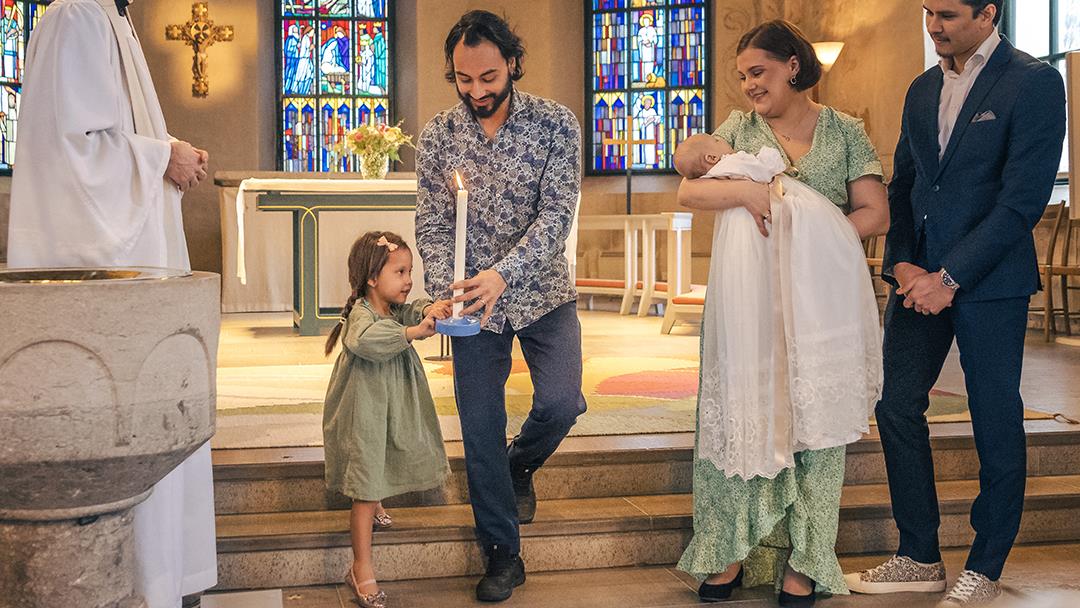 Ett dop i en kyrka. En liten flicka bär ett dopljus för att överlämna till det nydöpta barnet.