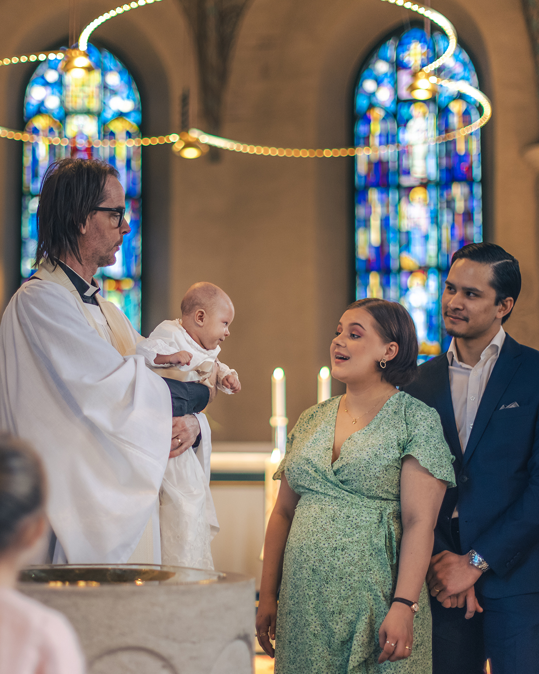 Ett dop i en kyrka. En kvinna och hennes man står bredvid prästen som håller barnet i famnen.