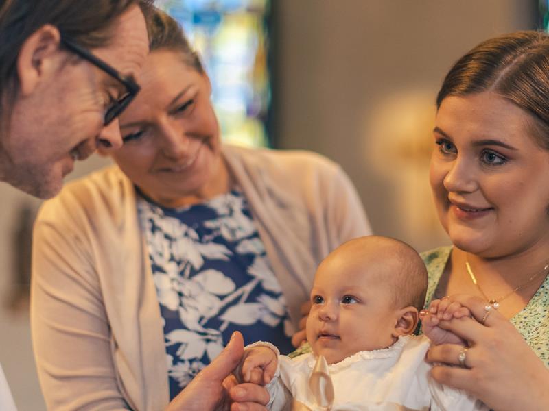 Ett dop i en kyrka. En kvinna med sin man bakom sig, håller barnet i famnen. En manlig präst håller barnets hand.