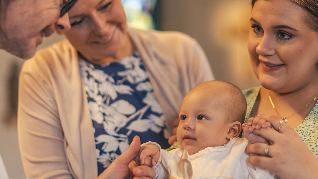 Ett dop i en kyrka. En kvinna med sin man bakom sig, håller barnet i famnen. En manlig präst håller barnets hand.