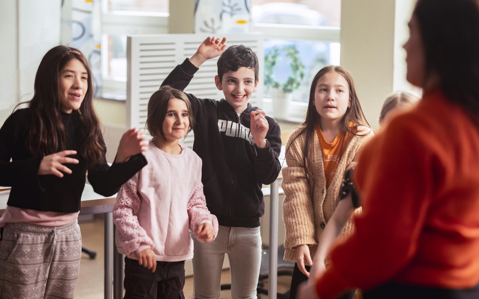 En barnkör sjunger och gestikulerar med armarna i ett klassrum. En kvinnlig körledare i förgrunden.