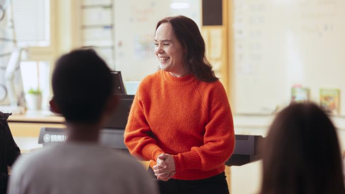 En kvinna i orange stickad tröja står och ler framför några barn i ett klassrum.