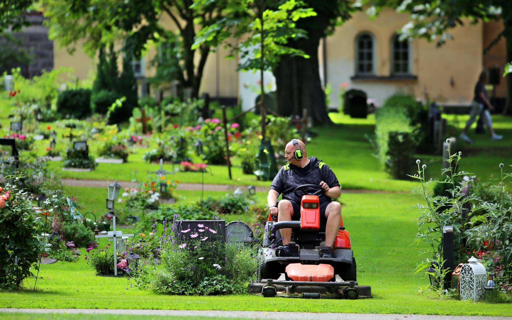 En kyrkogårdsarbetare klipper gräset runt gravstenarna med en åkgräsklippare.