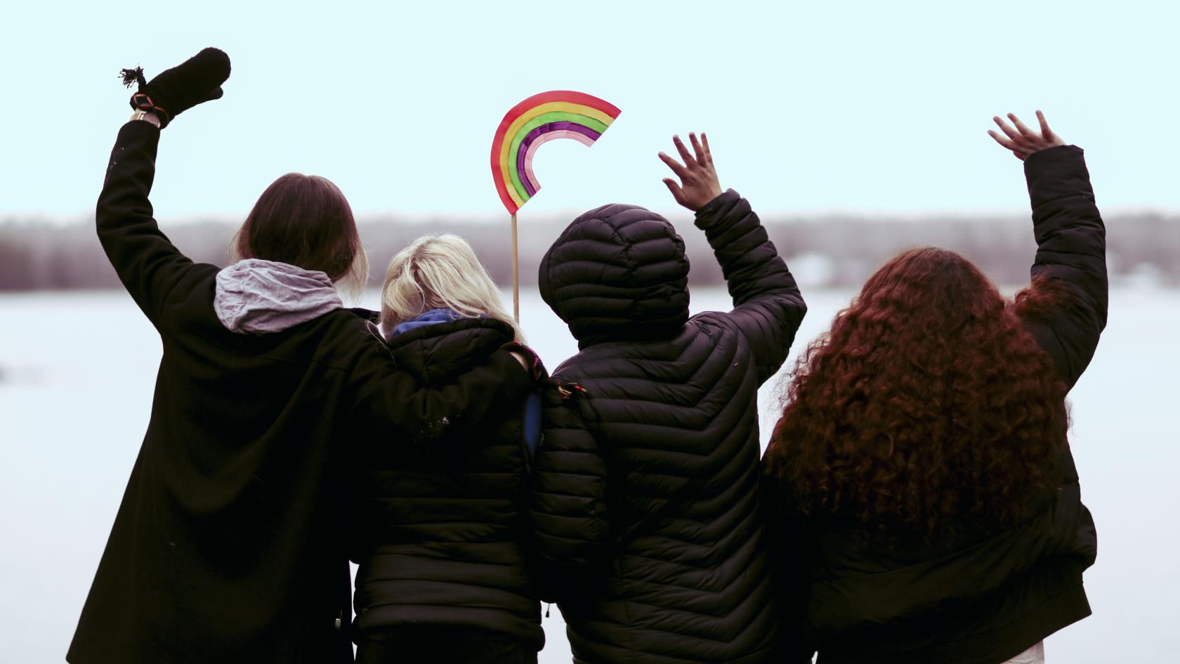 Fyra ungdomar står med ryggen mot kameran, vända mot vattnet och vinkar. En av dem håller i en regnbåge på en pinne.