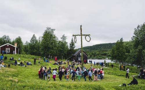 Ett midsommarfirande vid en sjö. Vuxna och barn dansar runt stången.