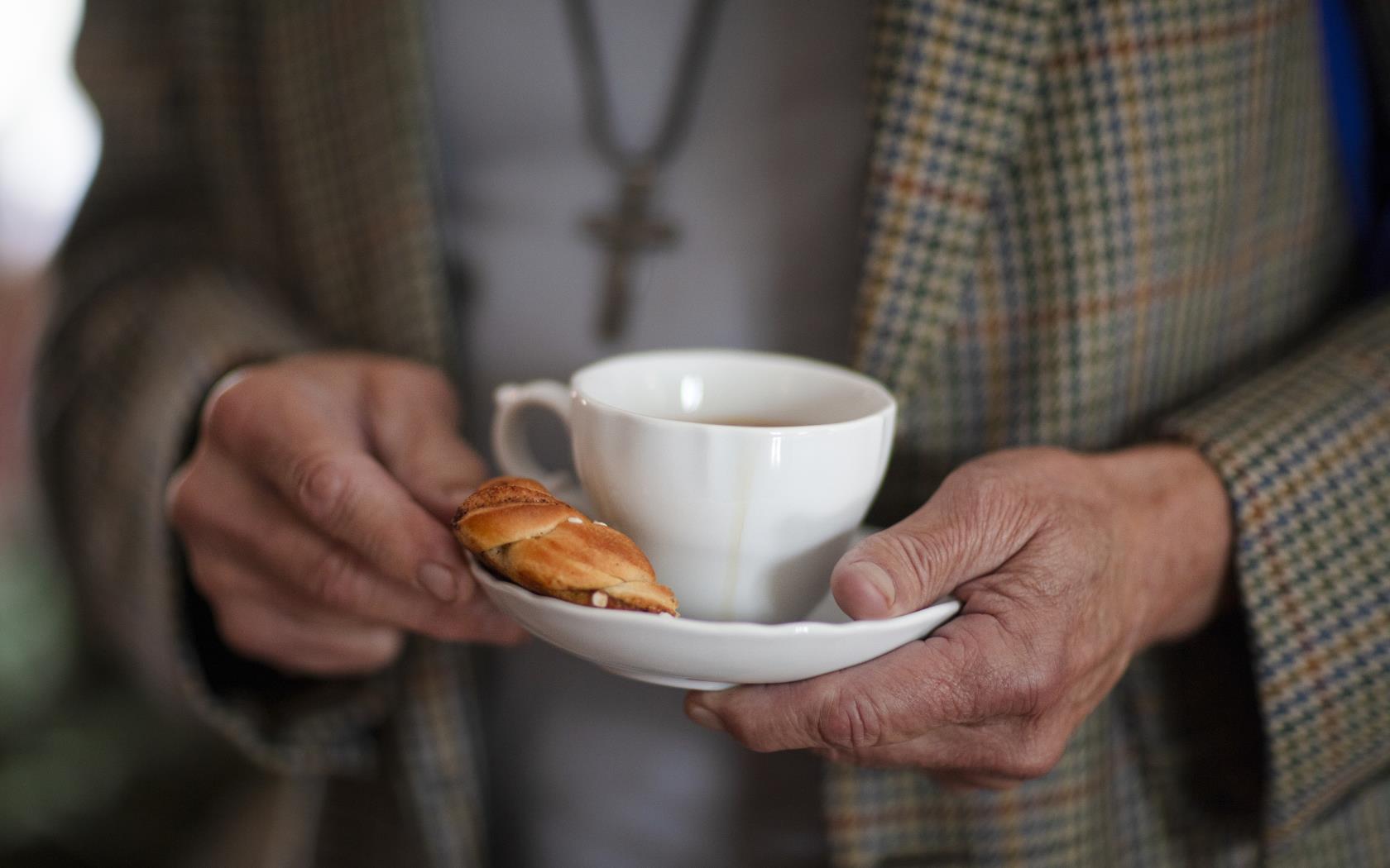 Någon i rutig kavaj håller en kaffekopp och en kanelbulle på fatet.