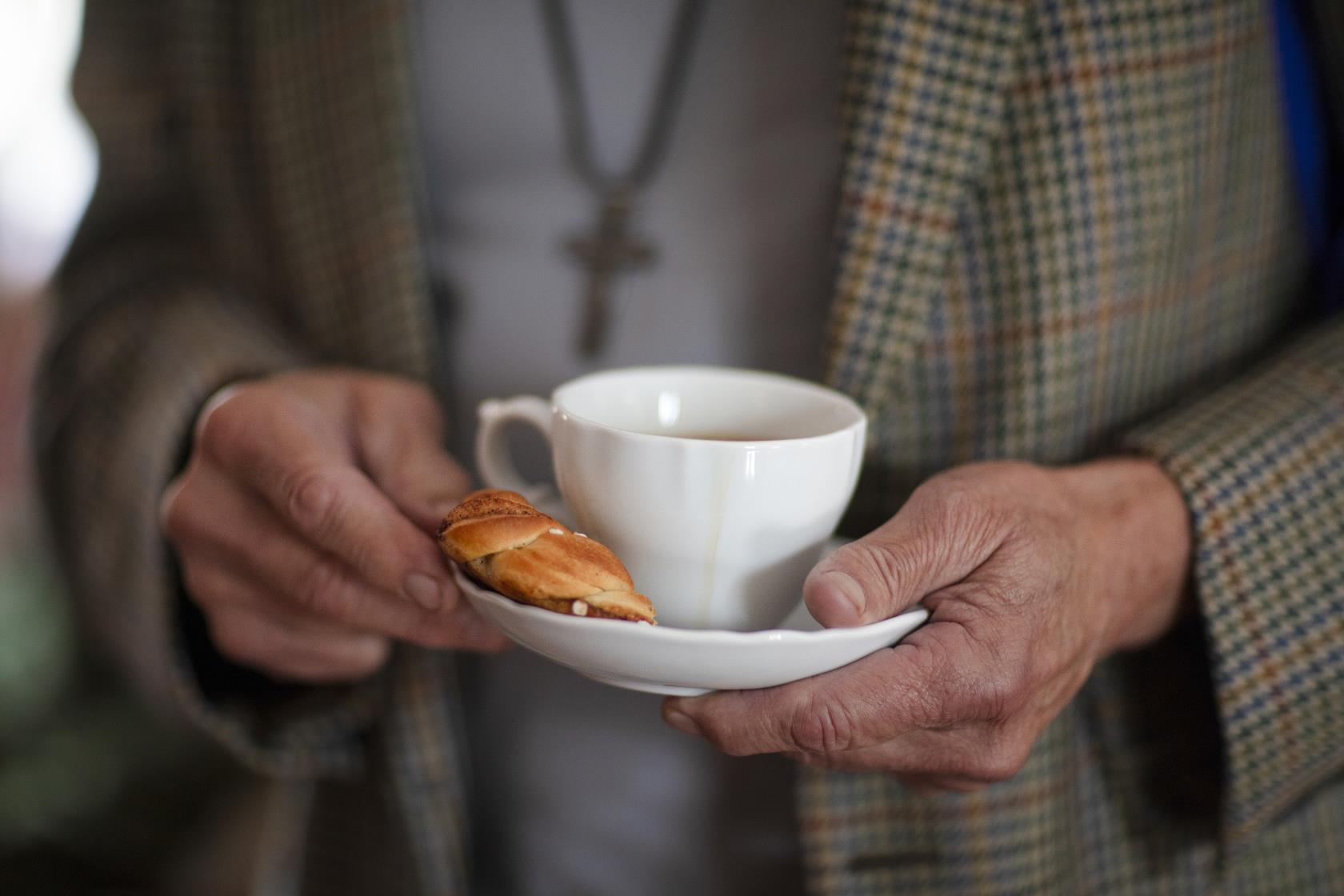 Någon i rutig kavaj håller en kaffekopp och en kanelbulle på fatet.