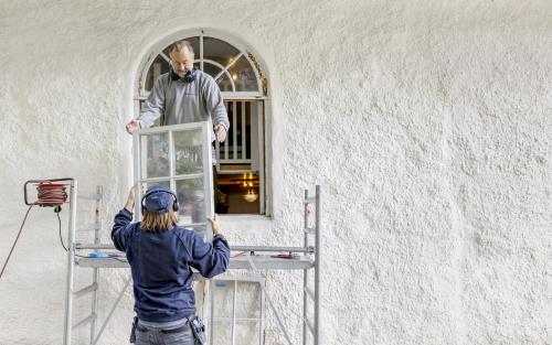 Två personer i arbetskläder monterar ner ett gammalt fönster.